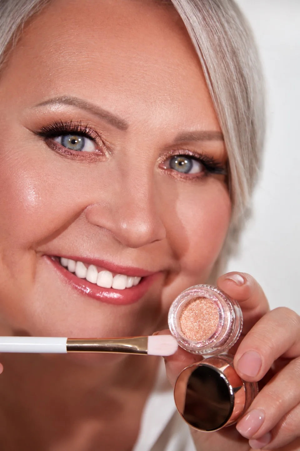 Smiling silver-haired woman holding a pink shimmer pigment jar and a makeup brush