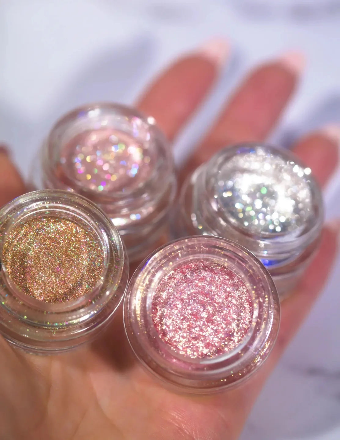 Four open shimmer pigment jars held in a hand, showing pink, gold and silver tones