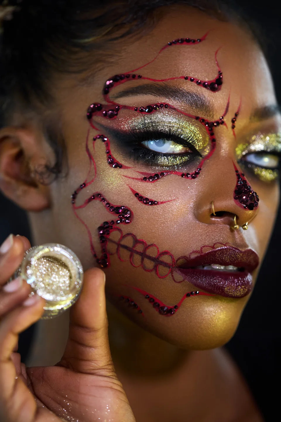 Gold and silver shimmer eyelids with burgundy jeweled skull lines and an open pigment jar