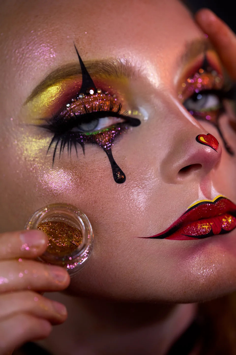 Pink and gold shimmer harlequin makeup with graphic black liner and a red heart nose