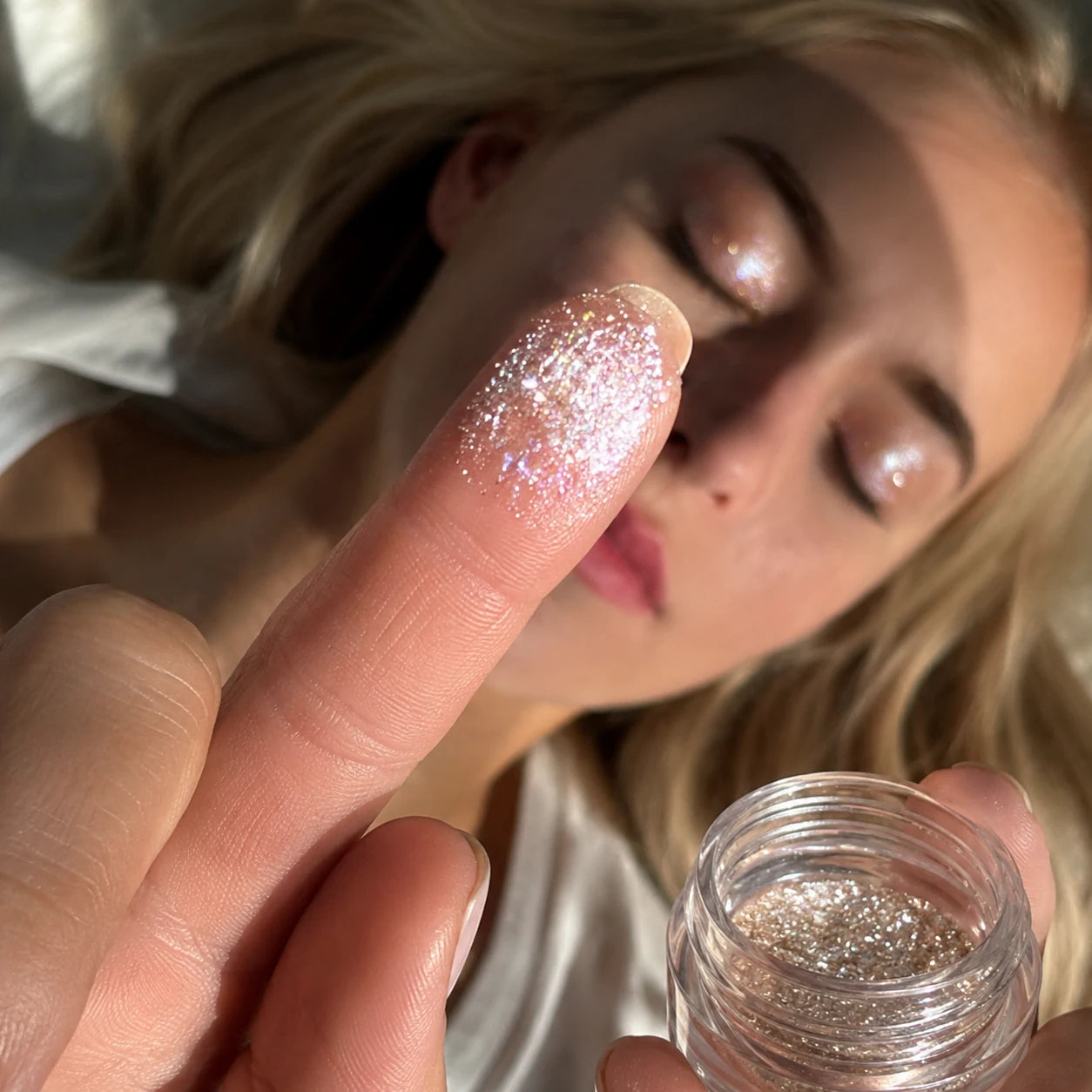 Close-up of intensely reflective loose pigment on a fingertip, with the open glass jar and shimmering eyelids behind it