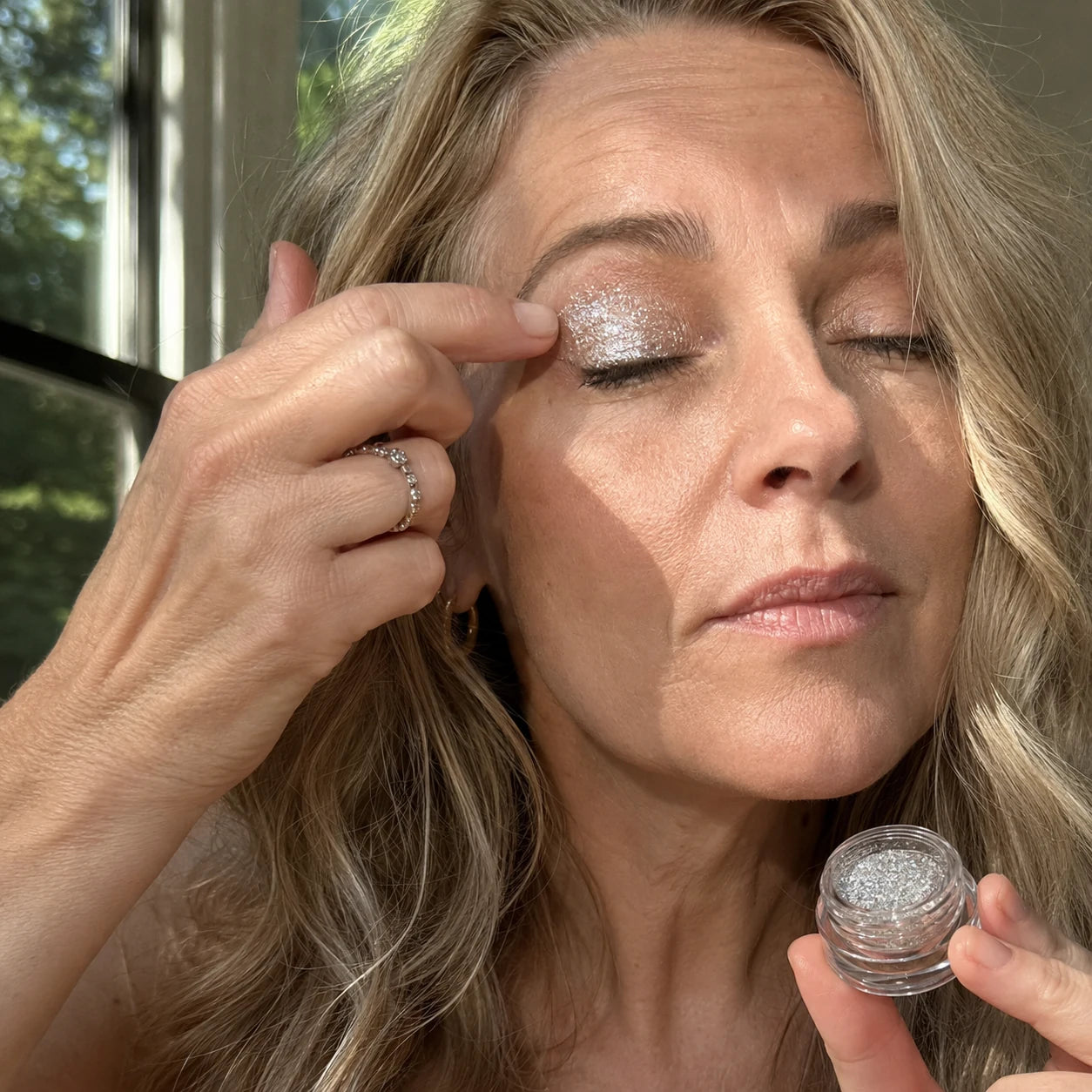 A mature blonde woman pressing silver shimmer onto her eyelid with a fingertip
